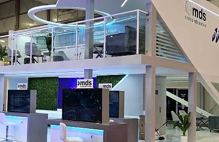 جناح معرض من طابقين لشركة mds System Integration، بشرفة علوية ذات حواجز زجاجية، وشاشات ومكاتب مضاءة في الأسفل