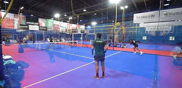 بطولة Padel In KSA: لاعبون على ملعب بادل داخلي أزرق تحت الكشافات، ولافتات العلامات التجارية حول الصالة