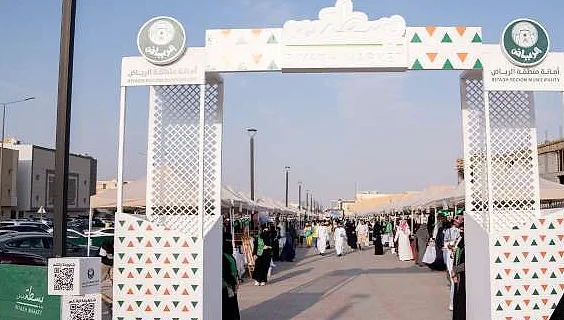 سوق الرياض: قوس مدخل أبيض شبكي بواجهة مزخرفة، ينفتح على ممر من أكشاك السوق والزوار