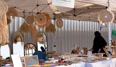 كشك حِرف يدوية في سوق الرياض تحت مظلة بيضاء، تتدلى منه قطع من القش المنسوج فوق طاولة من المصنوعات اليدوية