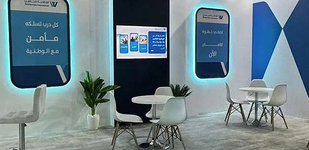 جناح الوطنية للتأمين: جدران بيضاء بألواح رسائل مضاءة من الخلف، وشاشة جدارية، وطاولات مقهى